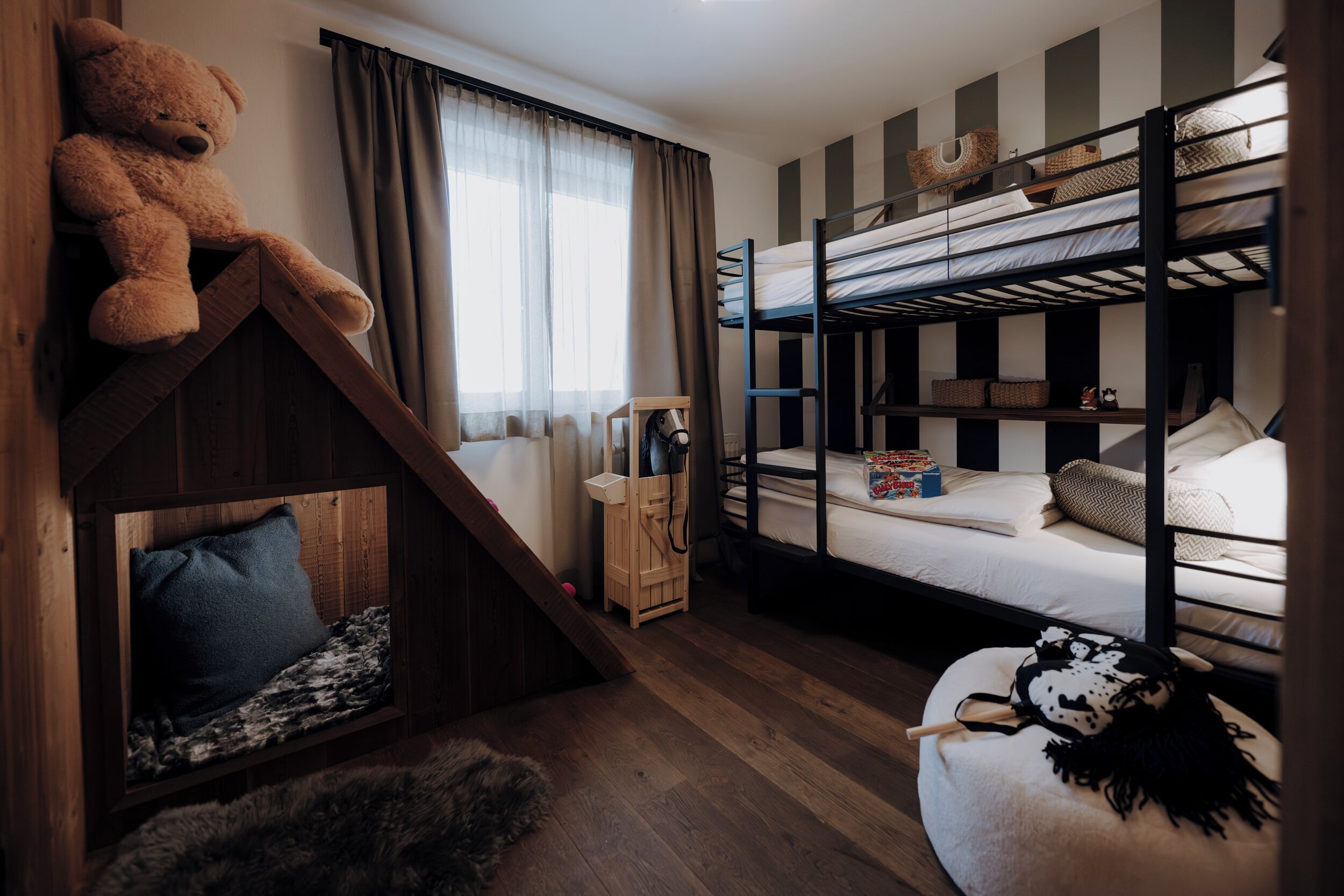 Habitación con literas, un oso de peluche, una pequeña escalera de madera y una ventana con cortinas.