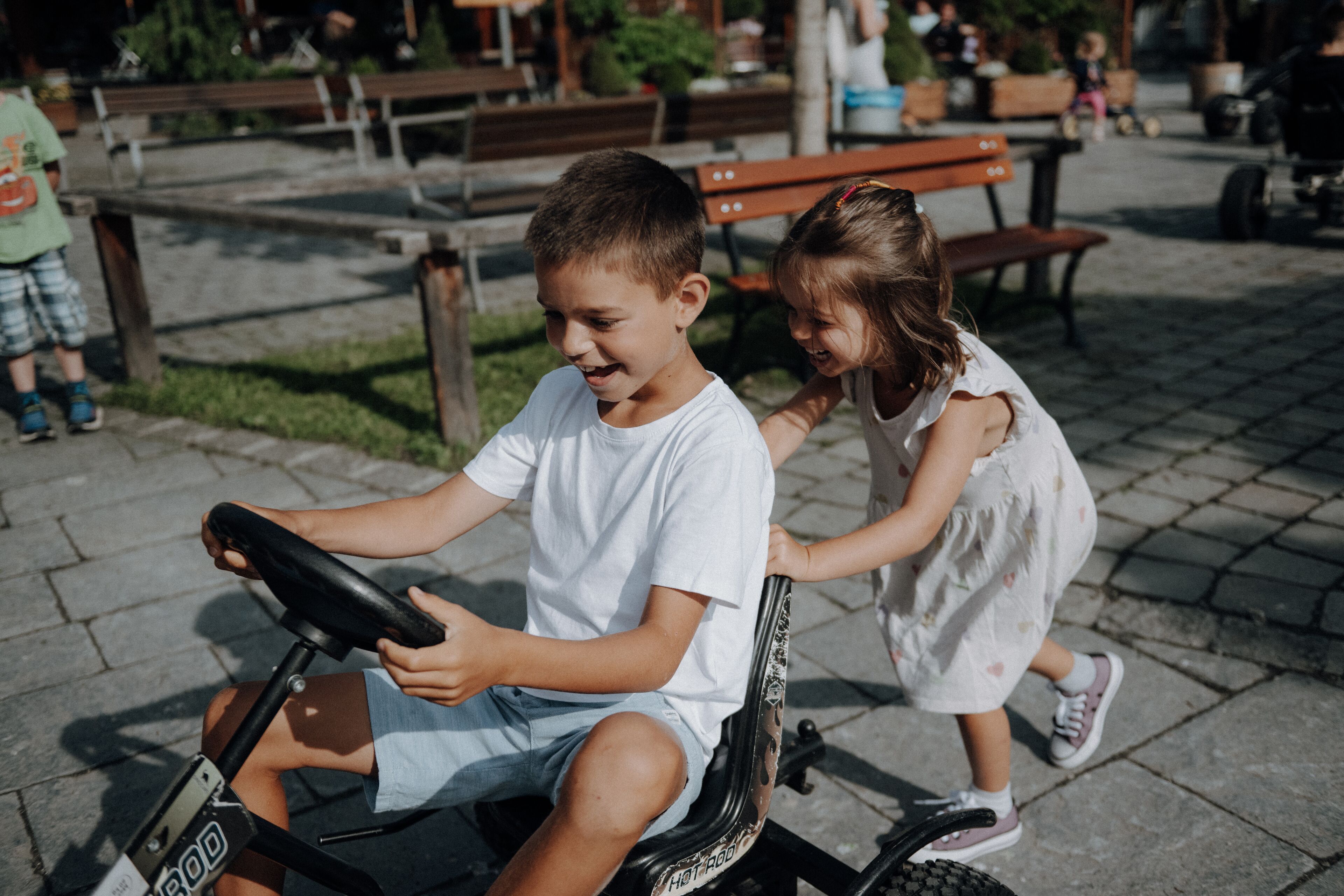 A child riding a small electric vehicle in a park with benches and greenery in the background.