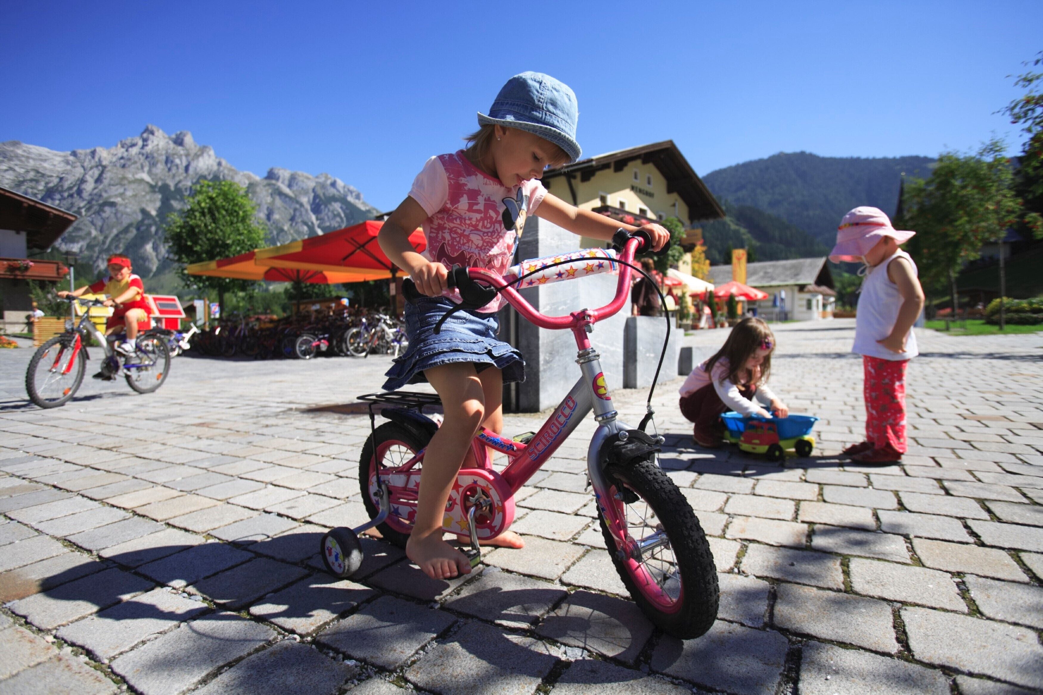 A child riding a pink bicycle on a paved area with mountains in the background.