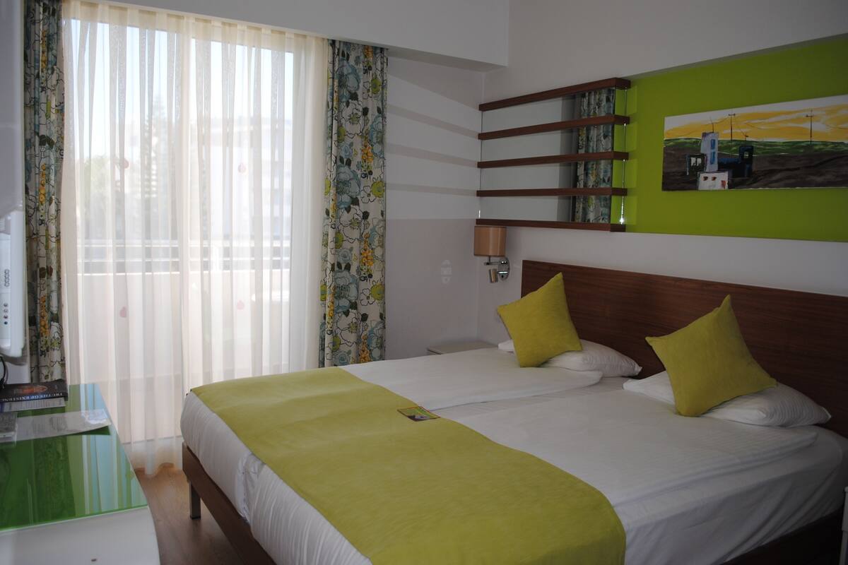 A hotel room with a large bed, a bedside table, a lamp, a window with curtains, and a painting on the wall.