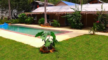 Une piscine avec une structure dotée d’un toit de chaume en arrière-plan, entourée d’une végétation luxuriante et d’une pelouse bien entretenue.
