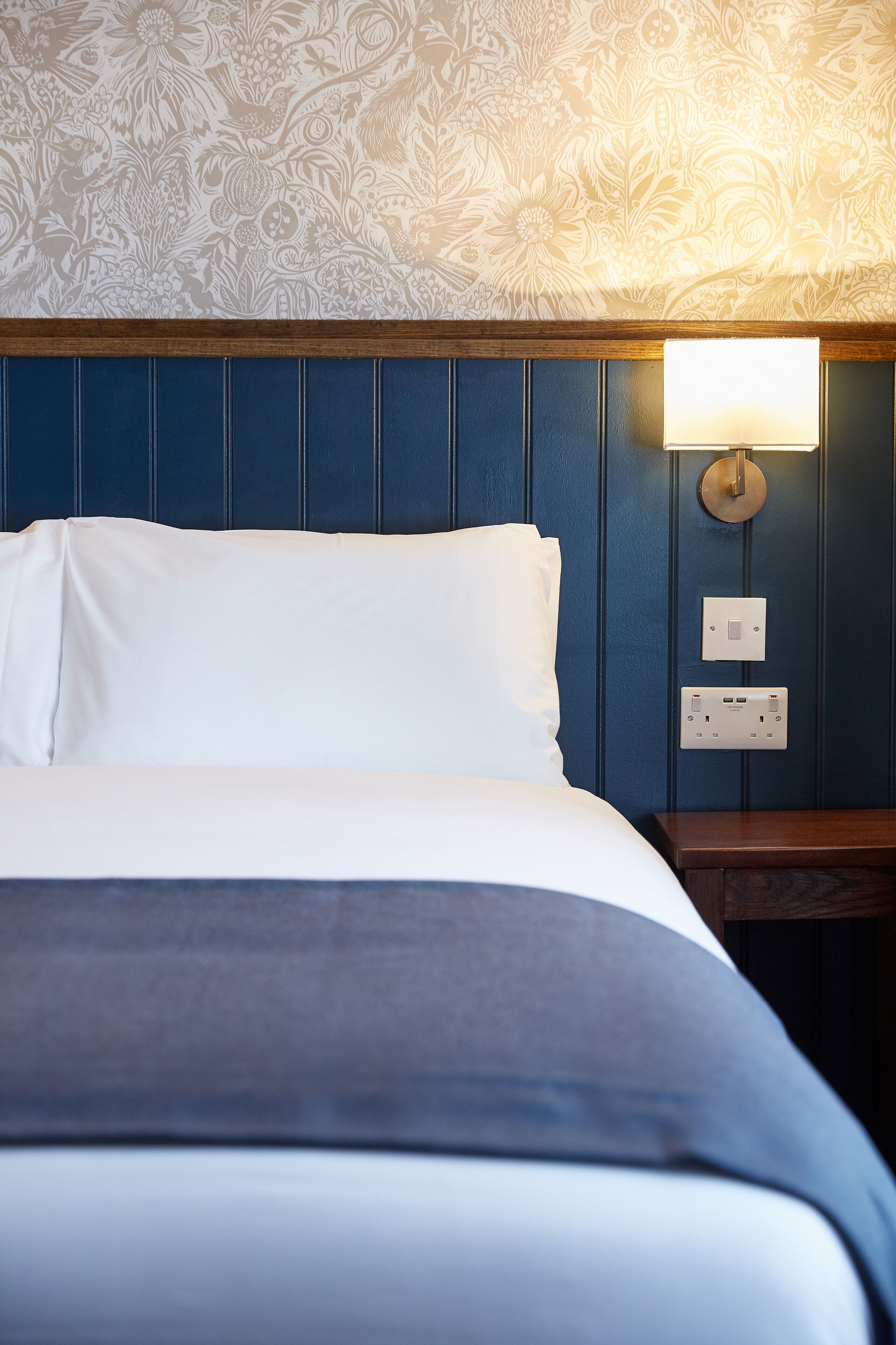 A neatly made bed with white linens and a dark blue headboard. A wall-mounted light fixture is visible to the right of the bed.