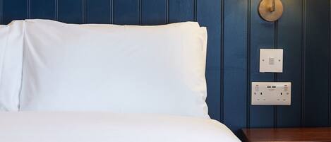 A neatly made bed with white linens and a dark blue headboard. A wall-mounted light fixture is visible to the right of the bed.