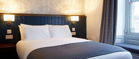 A neatly made bed with white and grey linens, a dark headboard, and two wall-mounted light fixtures.
