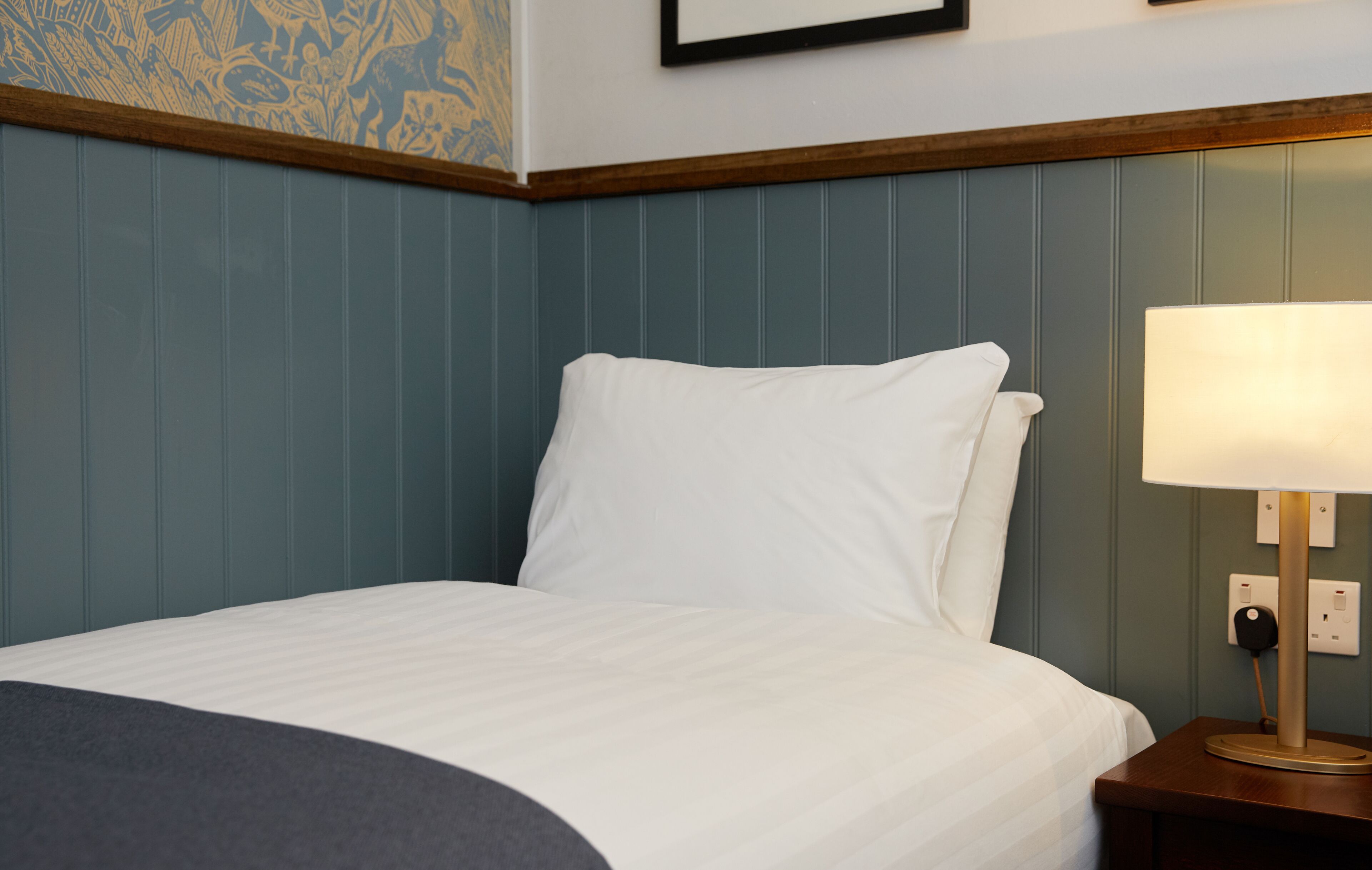 A neatly made bed with a white comforter and pillows, a wooden nightstand with a lamp, and a wall with a decorative panel.
