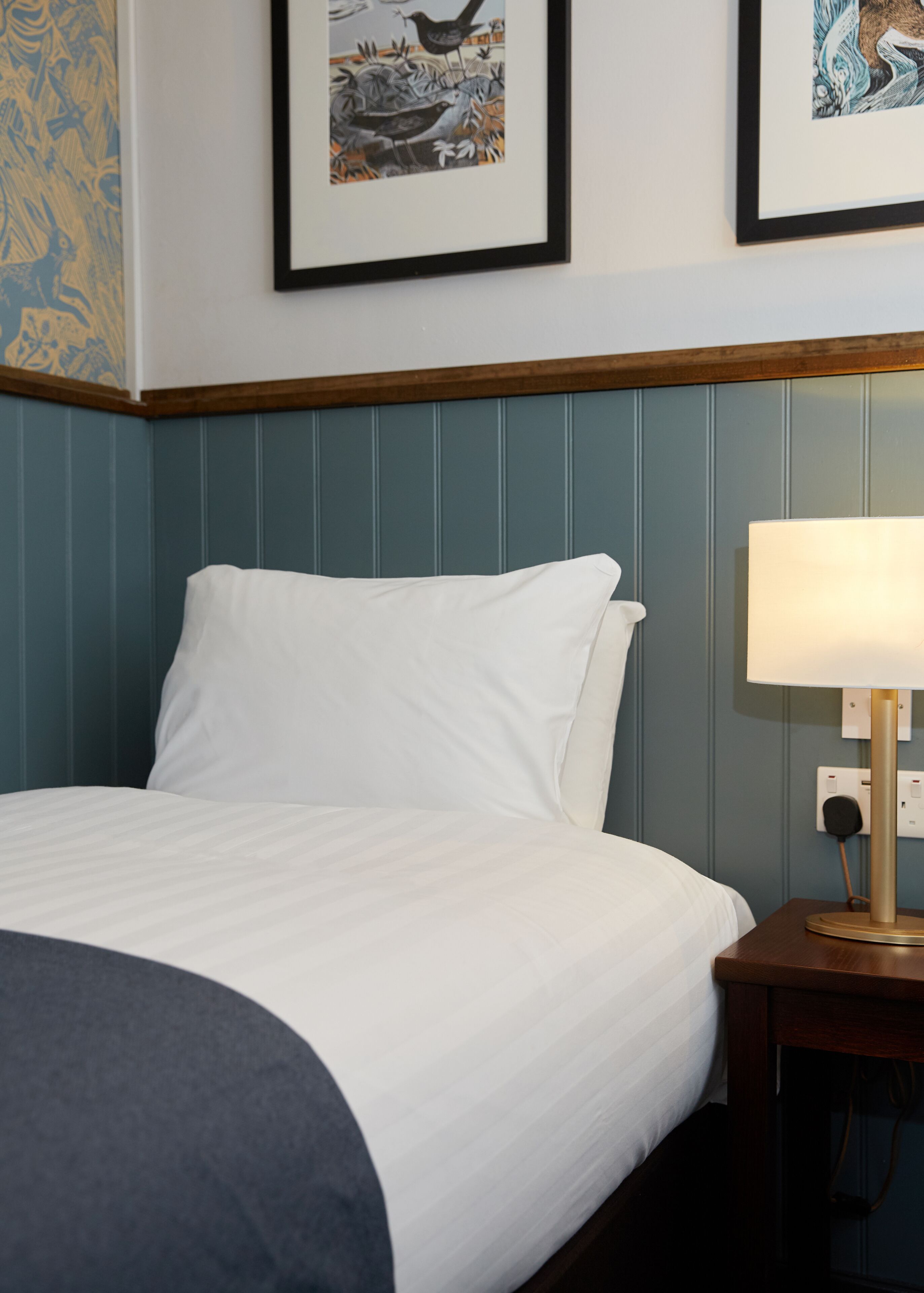 A neatly made bed with white linens, a wooden nightstand with a lamp, and two framed artworks on the wall.