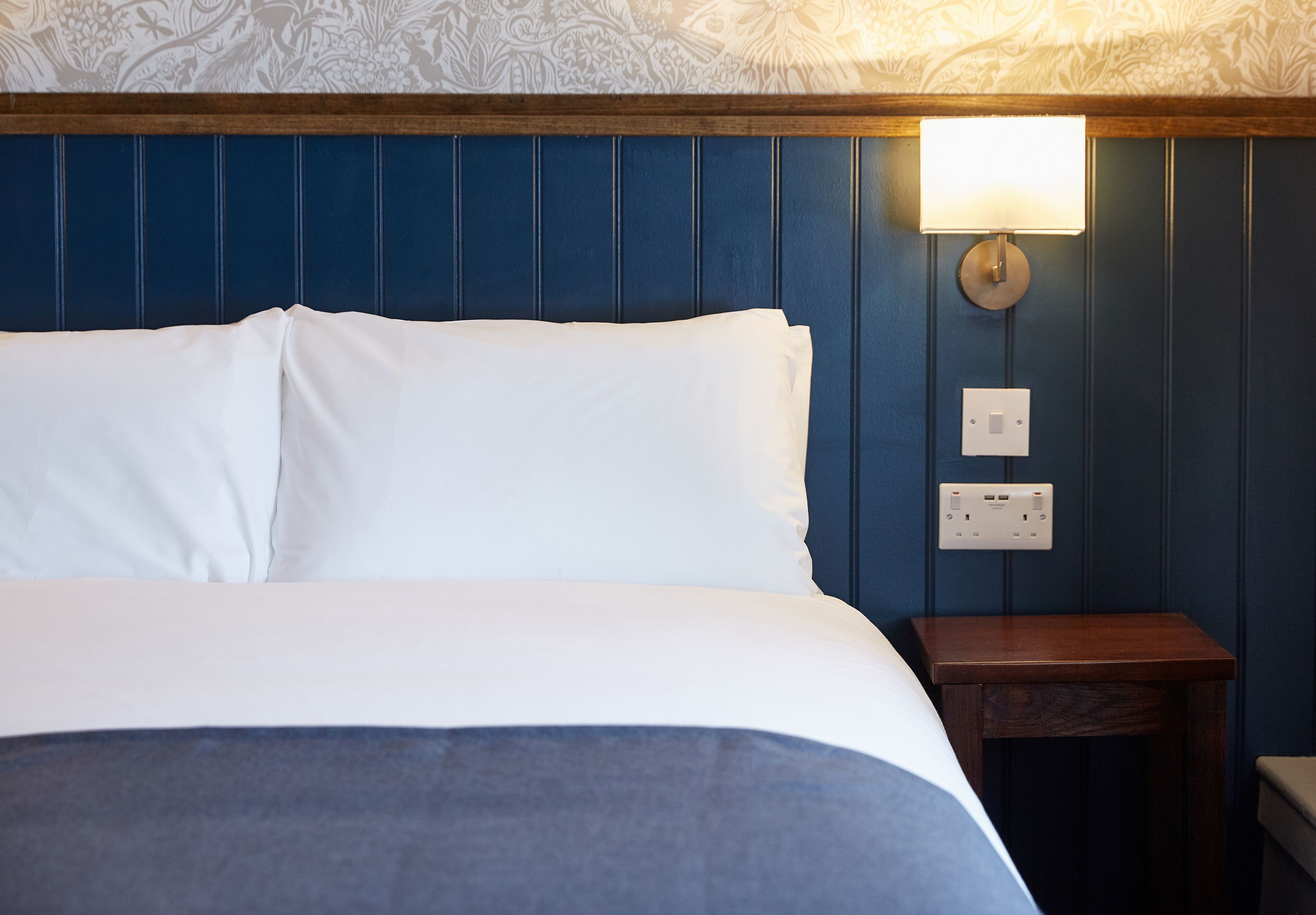 A neatly made bed with white linens, a wooden nightstand with a lamp, and a wall with a floral pattern.