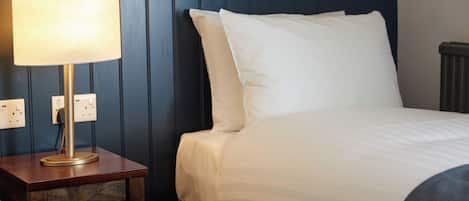 Una cama bien hecha con sábanas blancas y una manta a rayas, una lámpara de noche sobre una mesita de madera, y un muro azul con paneles.