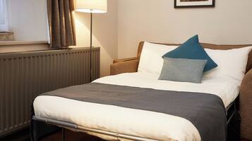 A neatly made bed with a grey blanket, a blue pillow, and a white pillow. A lamp is on the bedside table, and a framed picture hangs on the wall.