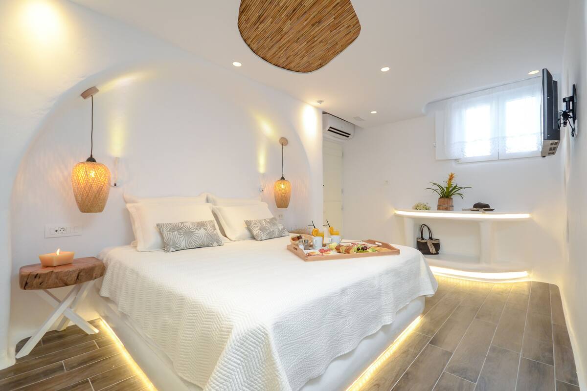 A modern bedroom with a large bed, bedside lamps, a tray of breakfast items, and a small shelf with toiletries.