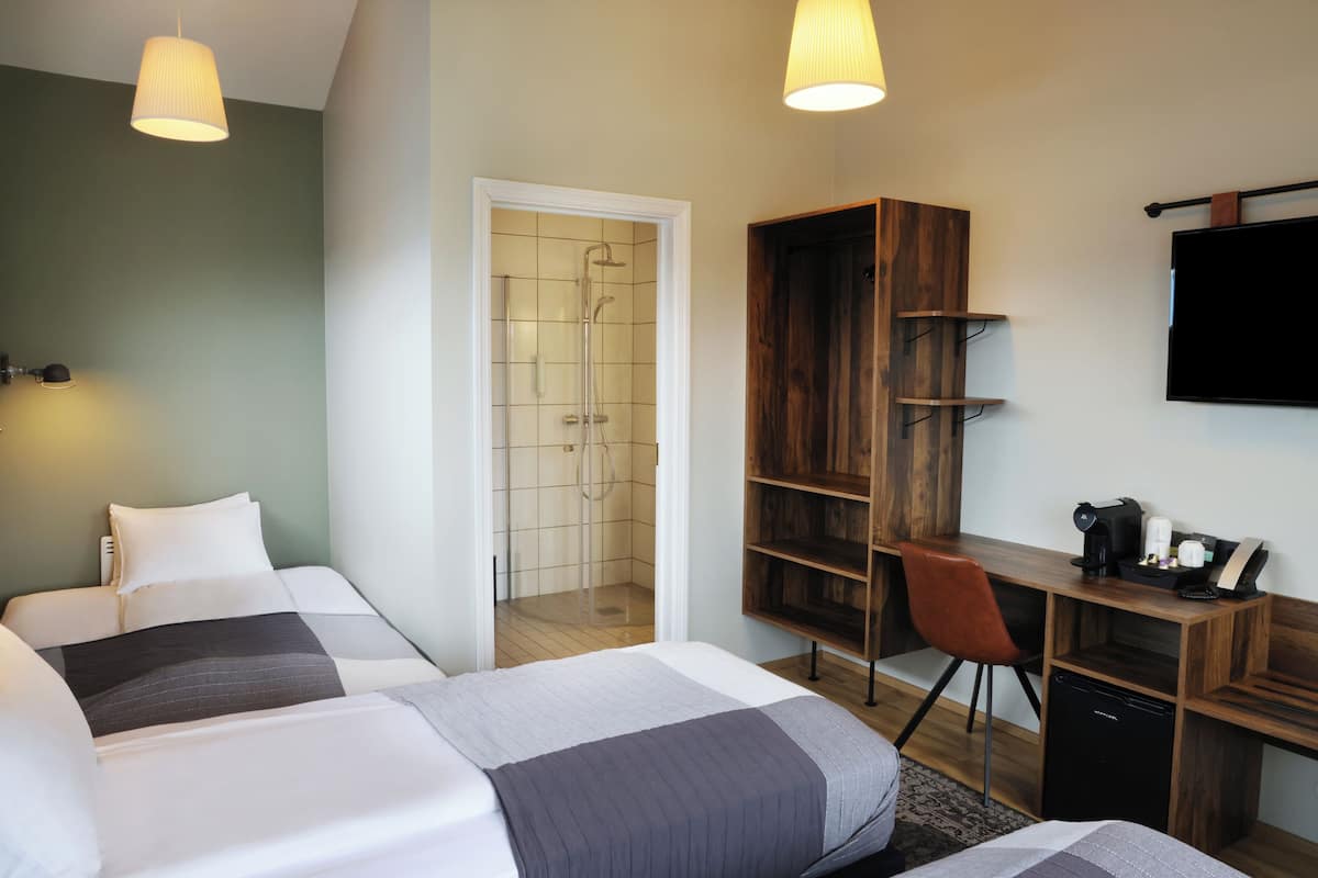 Ein Hotelzimmer mit zwei Betten, einem Schreibtisch mit Stuhl, einem Fernseher und einem Duschbereich.