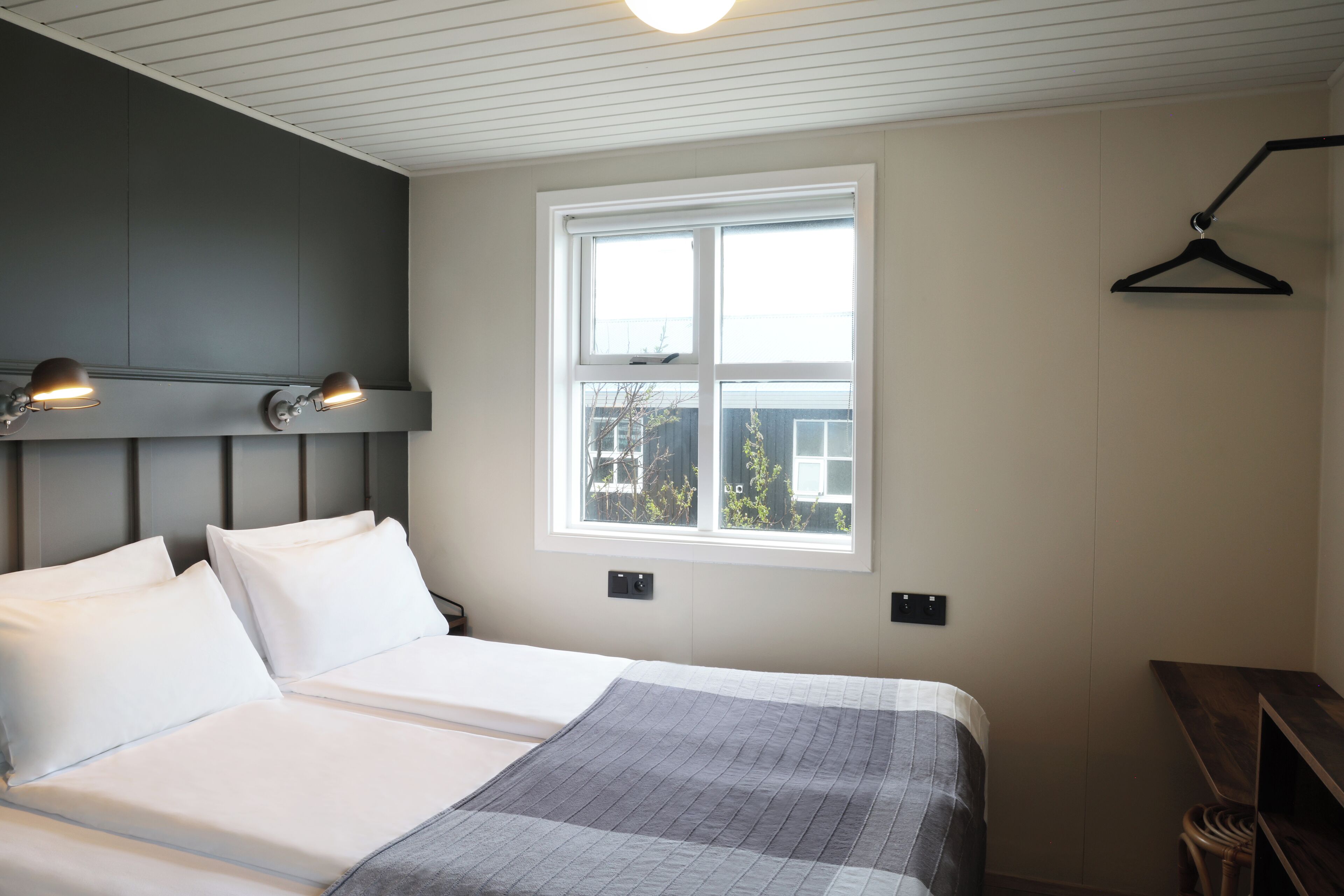 A neatly made bed with white linens and a gray blanket, a window with a view of trees, and a wall-mounted light fixture.