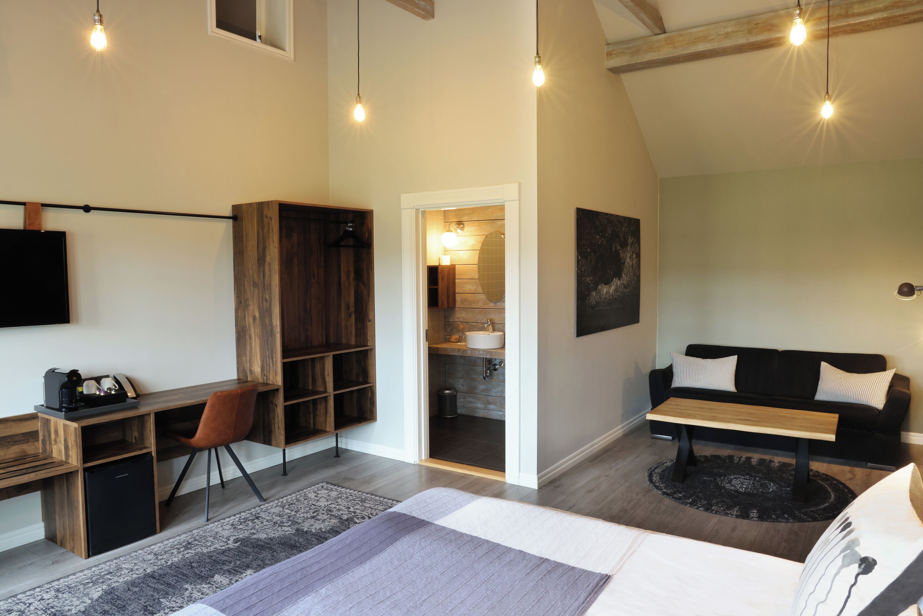 Una habitación moderna con un escritorio de madera, un sofá negro, una cama y un baño visible a través de una puerta abierta.