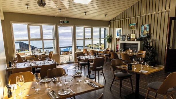 A restaurant with tables set for dining, large windows offering a view of the sea, and a fireplace on the wall. - Fosshotel Hellnar (Hellnar)