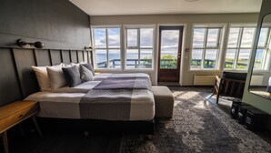 A bedroom with a large bed, a nightstand, a bench, and a view of the outdoors through large windows. - Fosshotel Hellnar (Hellnar)
