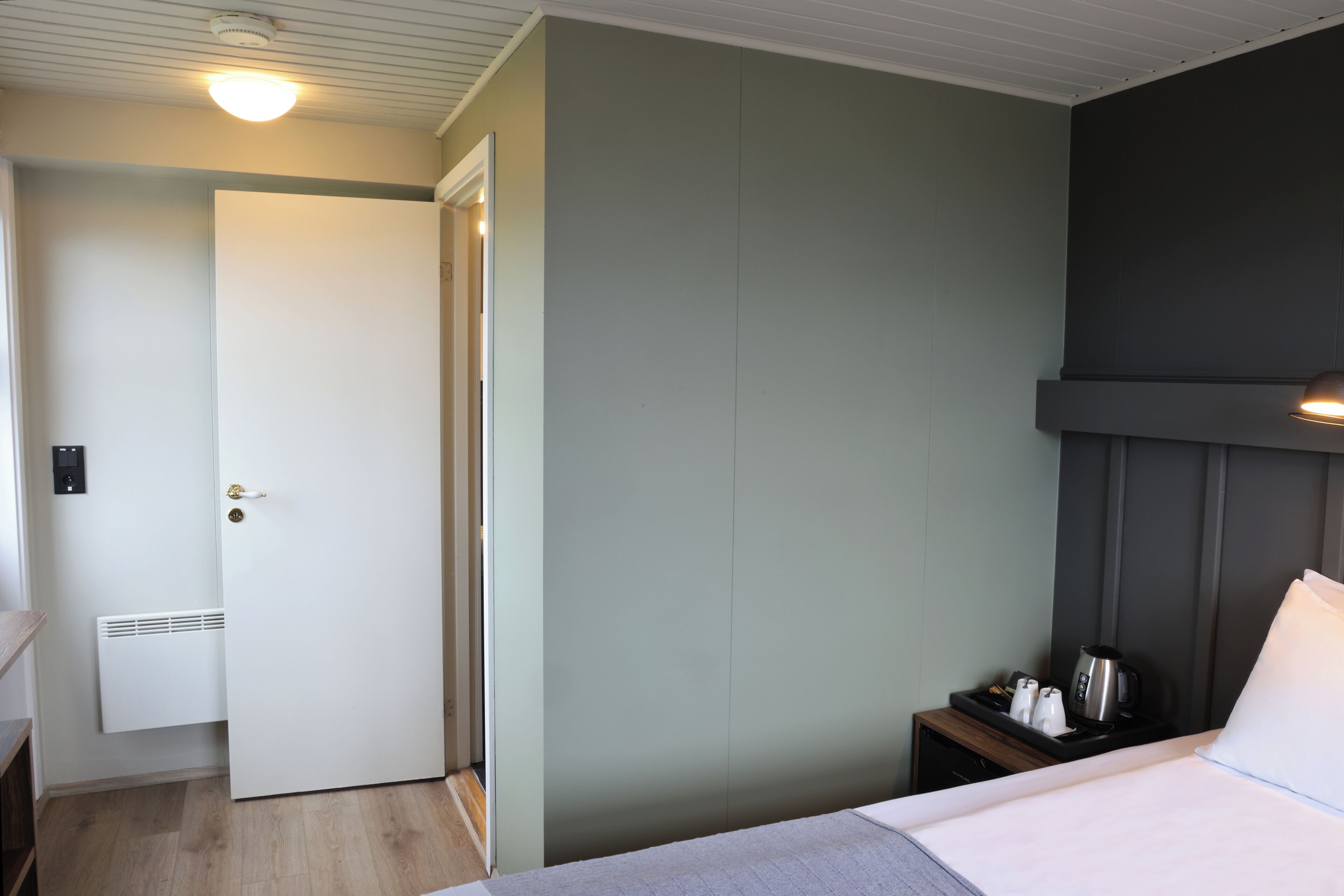 Una camera d'albergo con un letto, un comodino con bollitore e tazze, una porta e un termosifone a parete.