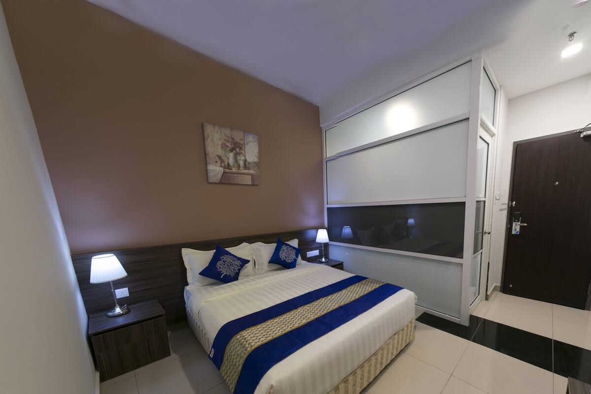 Une chambre d’hôtel avec un grand lit, deux tables de chevet avec des lampes, une fenêtre avec des stores, une porte et un tableau au mur.