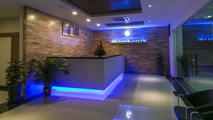 Uma área de recepção de hotel com design moderno, apresentando um balcão de atendimento, plantas em vasos e uma placa com a inscrição "Lavana Hotel".
