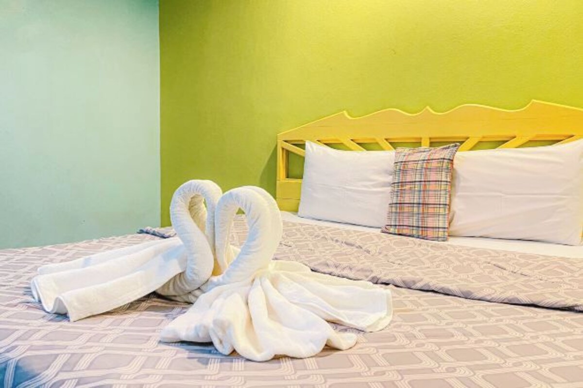 A bed with a yellow headboard, a plaid pillow, and two towels arranged in a swan shape.
