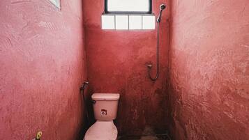A small, narrow bathroom with pink walls, a toilet, a window, and a shower.