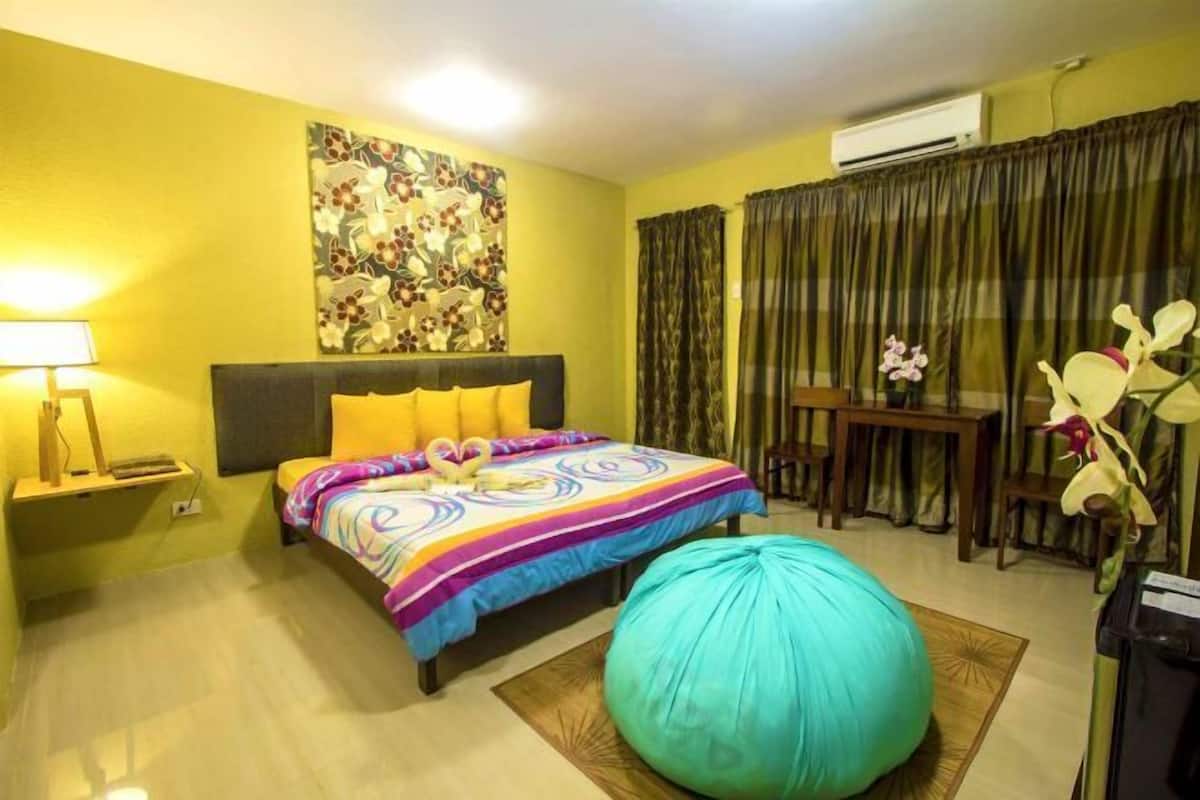 A hotel room with a bed, a desk, a chair, a teal bean bag, a wall-mounted air conditioner, and a floral artwork.