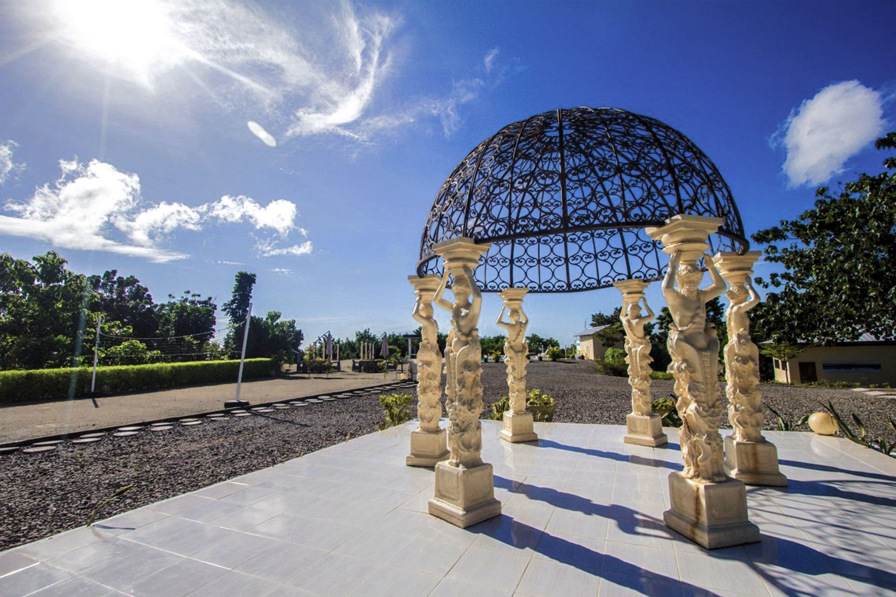 A gazebo with a dome and ornate columns, surrounded by a paved area and greenery.