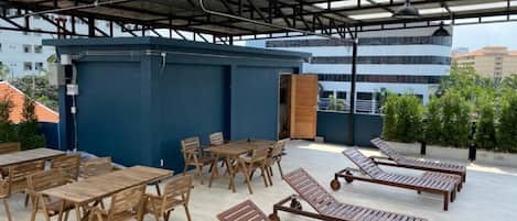 A rooftop patio with wooden tables and lounge chairs, a blue structure, and a metal roof.