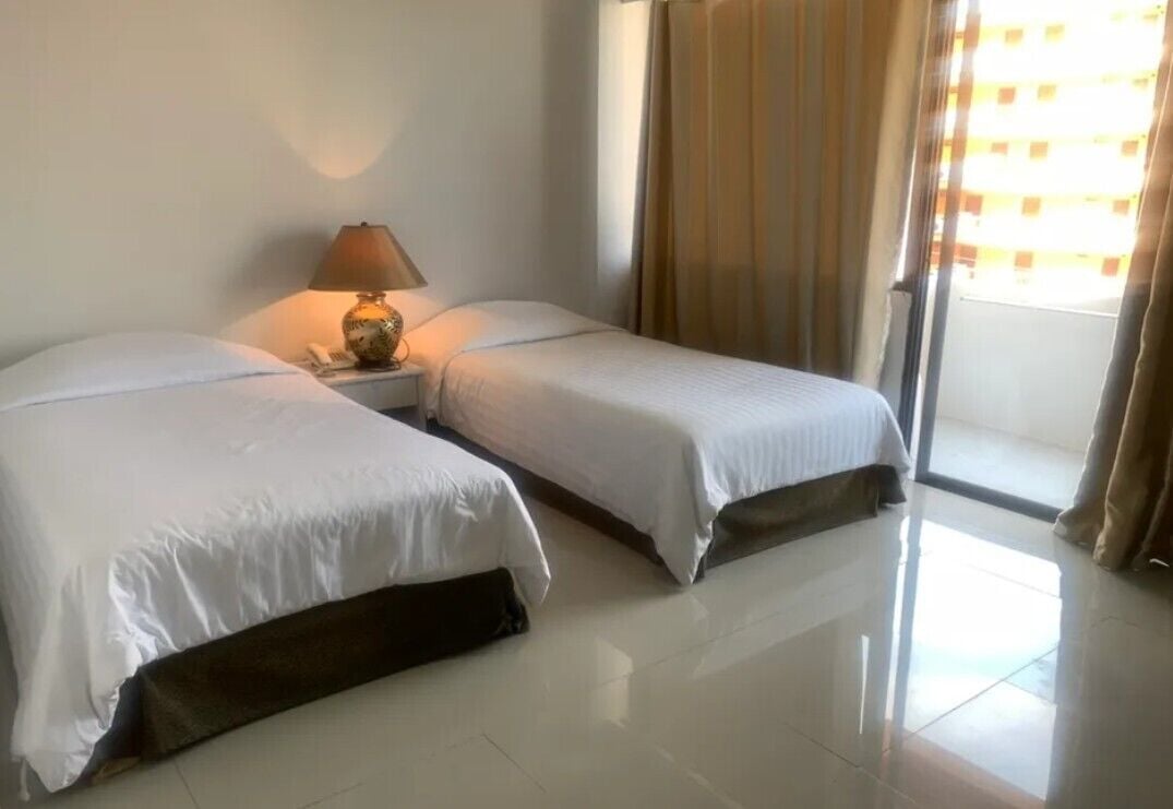 Une chambre d’hôtel avec deux lits, une table de chevet avec une lampe, une fenêtre avec des rideaux et une vue sur l’extérieur.