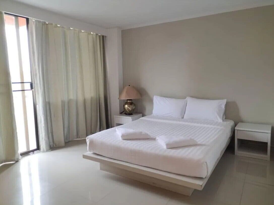 Een moderne hotelkamer met een groot bed, een nachtkastje met een lamp en uitzicht op het omliggende landschap door een raam met gordijnen.