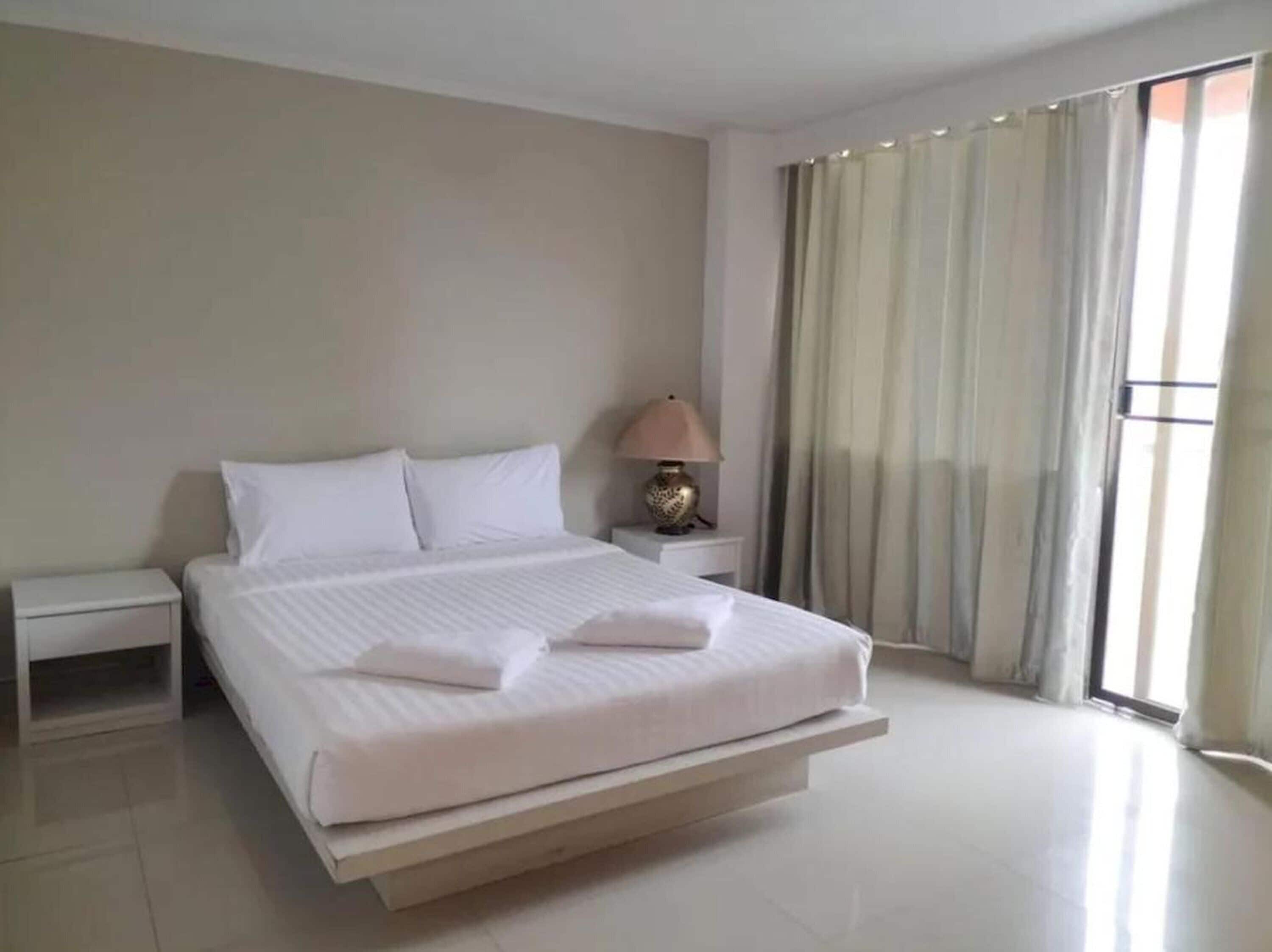 A modern hotel room with a large bed, a bedside table, a lamp, and a view of the outdoors through a window.