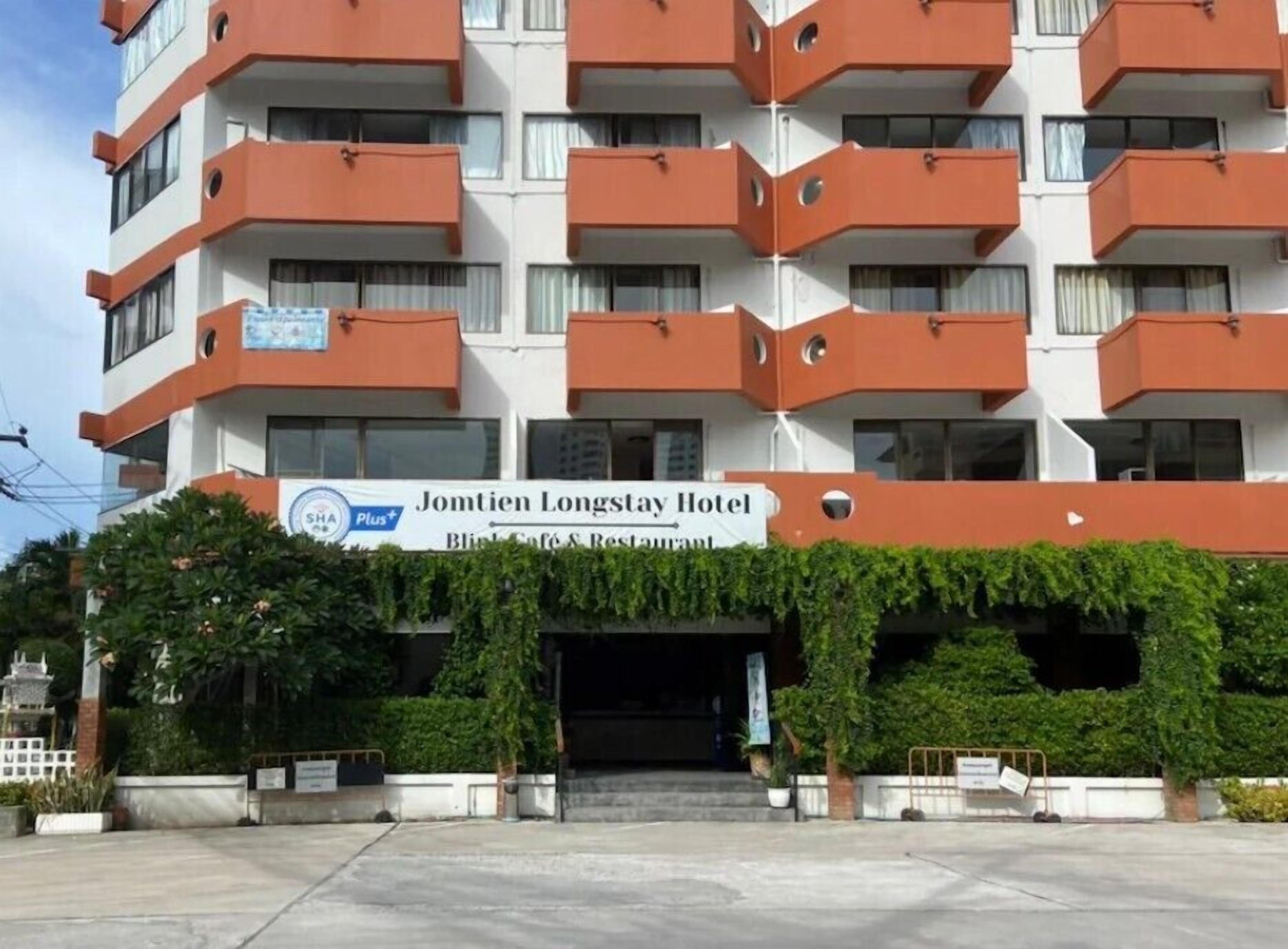 Um hotel com vários andares, varandas, uma placa com o nome Jomtion Longstay Hotel e uma pequena área de entrada com plantas.