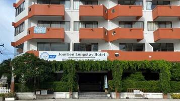 A multi-story hotel with balconies, a sign for the Jomtion Longstay Hotel, and a small entrance area with greenery.