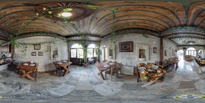Un comedor espacioso con altos techos de madera, paredes de piedra y ventanas amplias que dejan entrar la luz natural. Mesas dispuestas para una comida, y plantas en macetas que añaden verdor.