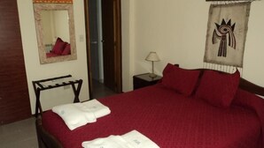 A neatly made bed with red bedding and white pillows, a wooden chair, a small bedside table with a lamp, and a wall tapestry. - Apart Hotel Tierra del Fuego (Ushuaia)