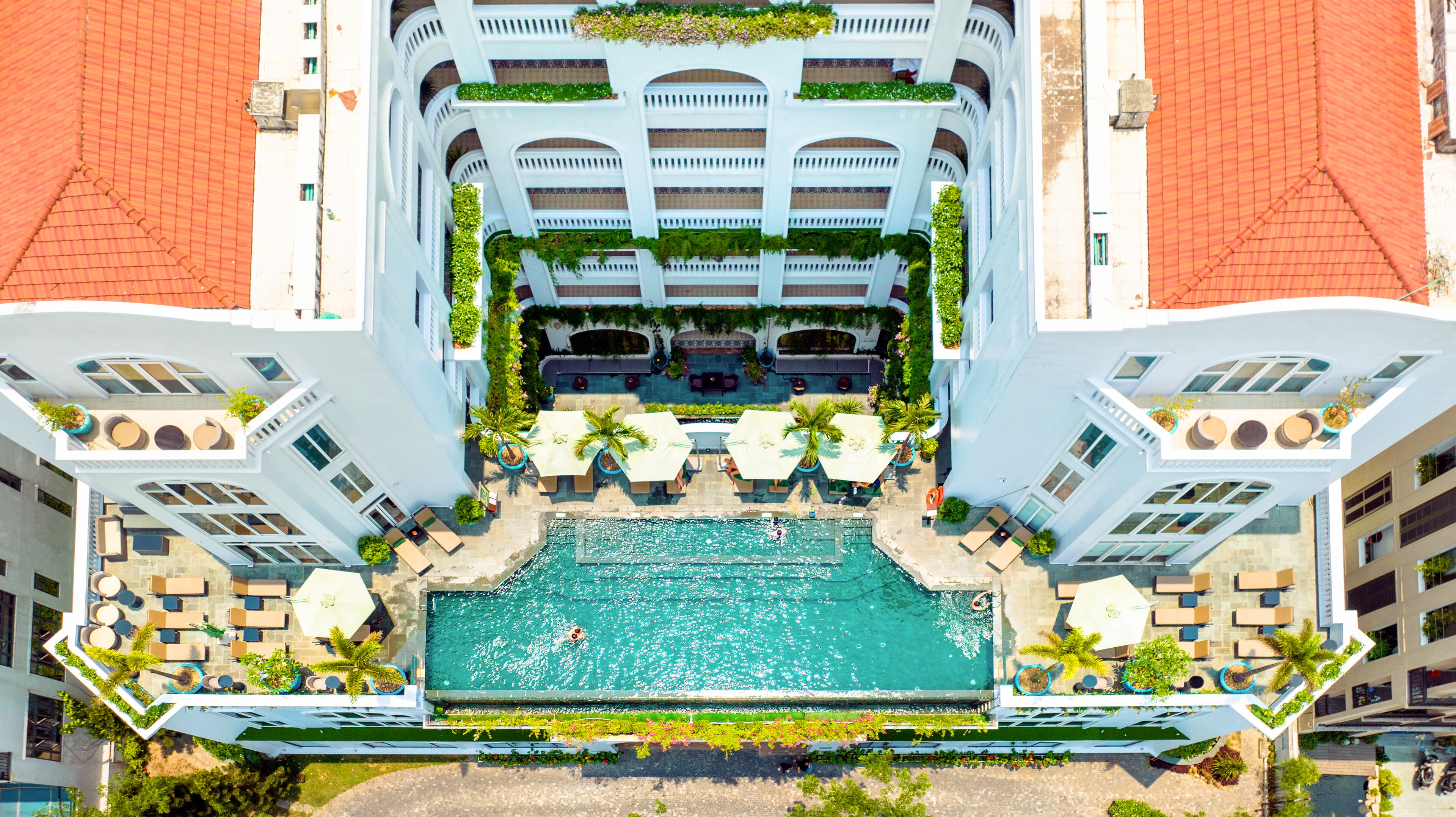 Vista aerea di un hotel con piscina centrale circondata da aree lounge all'aperto e vegetazione.