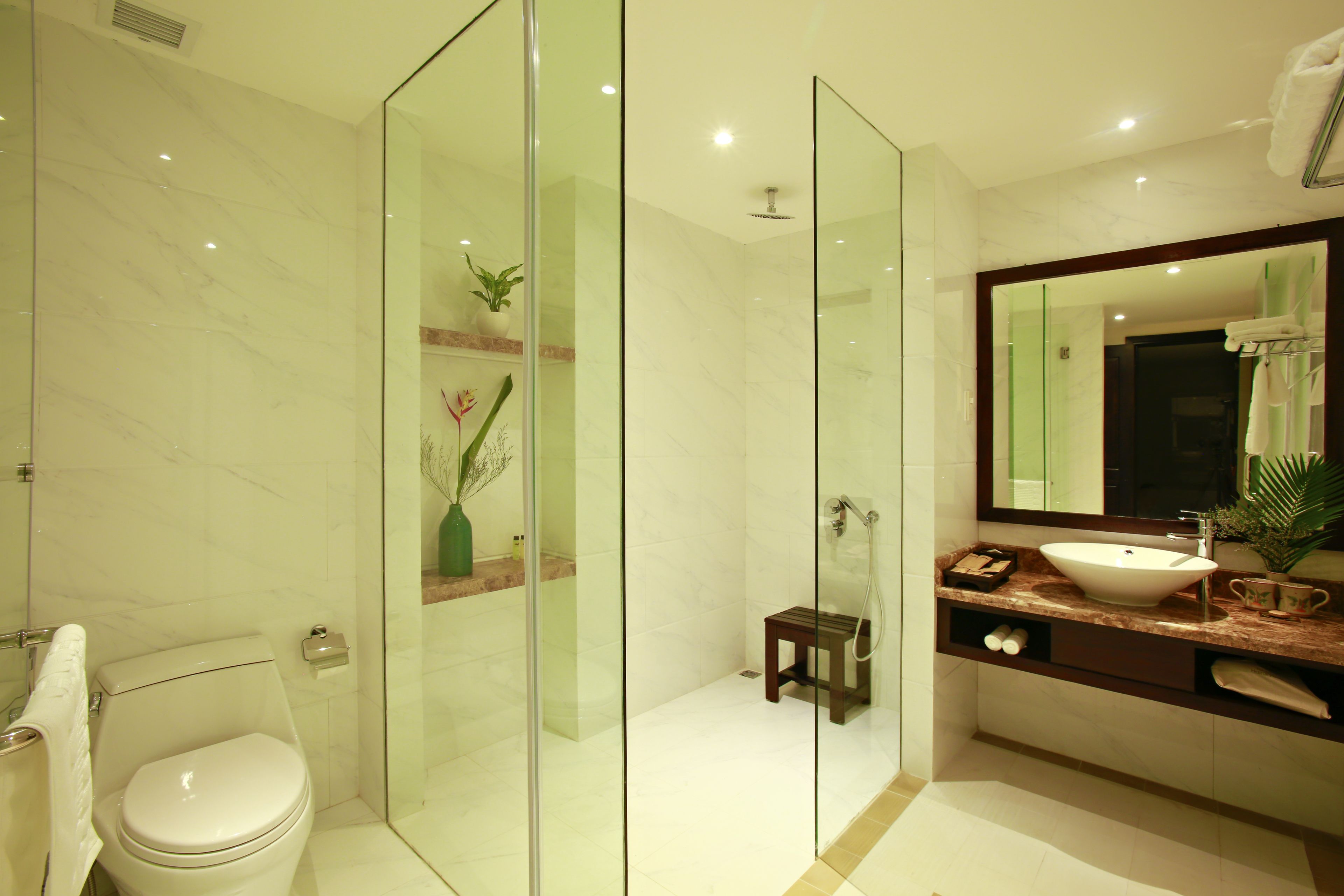 A modern bathroom with a glass-enclosed shower, a marble-tiled floor, a white toilet, a wooden vanity with a sink, and a large mirror.