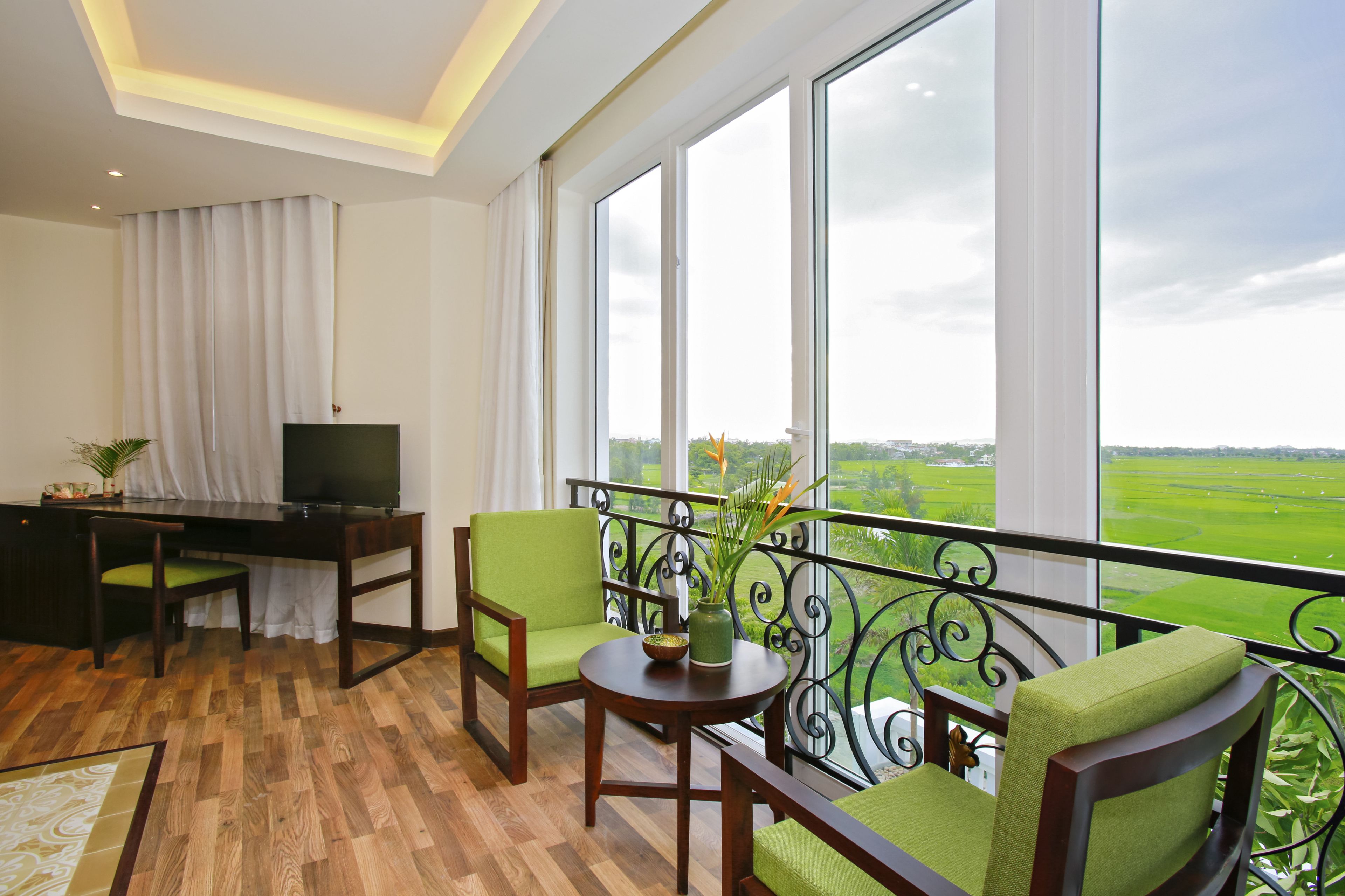 A balcony with a view of a green landscape, a desk with a television, and green upholstered chairs.