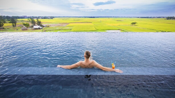 A man relaxing in a infinity pool with a view of green fields and a distant village. - Lasenta Boutique Hotel Hoian (Da Nang)