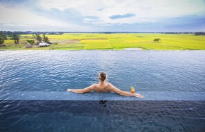 A man relaxing in a infinity pool with a view of green fields and a distant village.