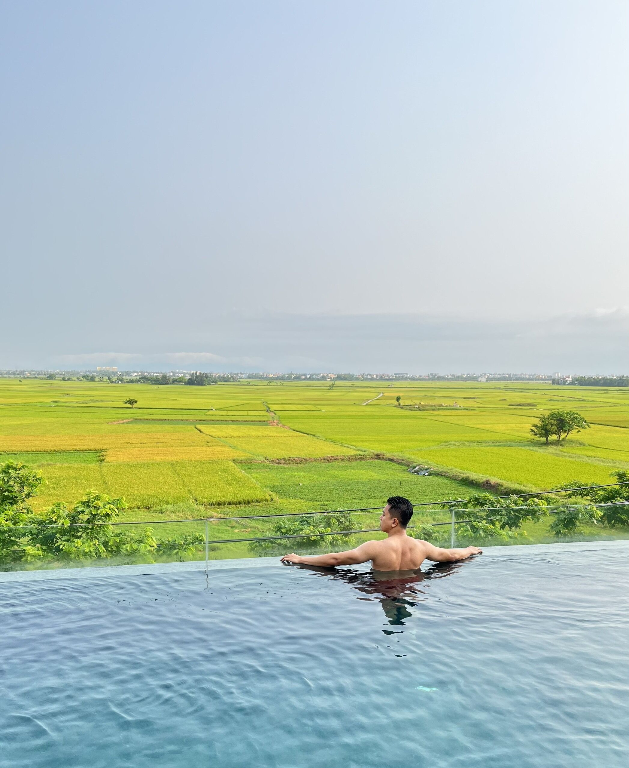A man relaxing in a infinity pool with a view of green fields and distant buildings.