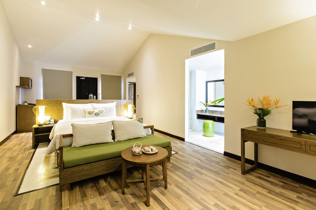 Une chambre d’hôtel moderne avec un lit en bois, un canapé vert, une petite table avec un plateau, un téléviseur posé sur un meuble en bois et une plante en pot.