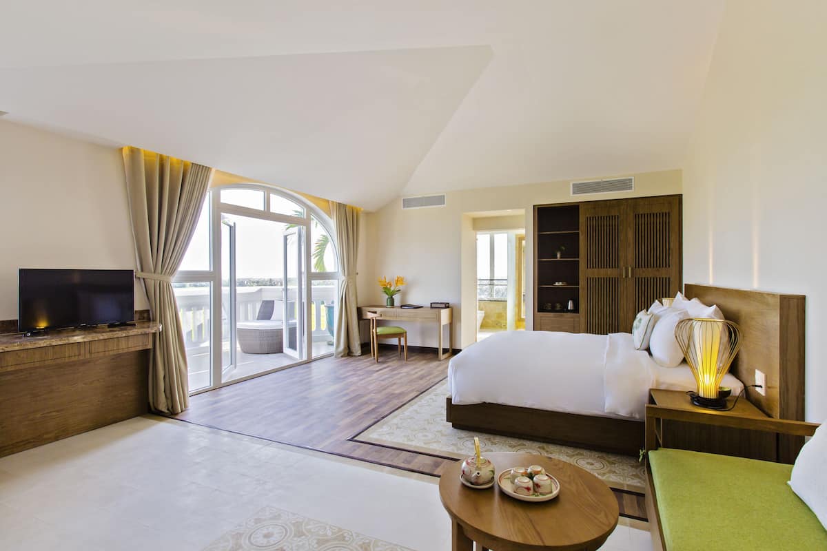 Une chambre d’hôtel comprenant un lit, un bureau, une télévision, une table à manger avec une théière, et un balcon avec des rideaux.