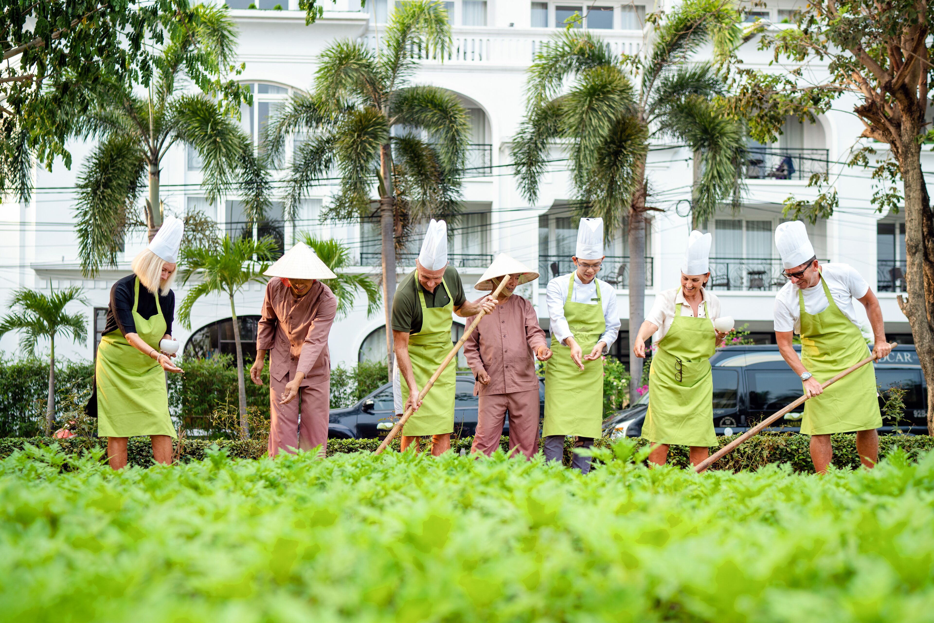 Menschen in Schürzen und Kochmützen arbeiten in einem Gartenbereich, im Hintergrund ist ein Gebäude zu sehen.