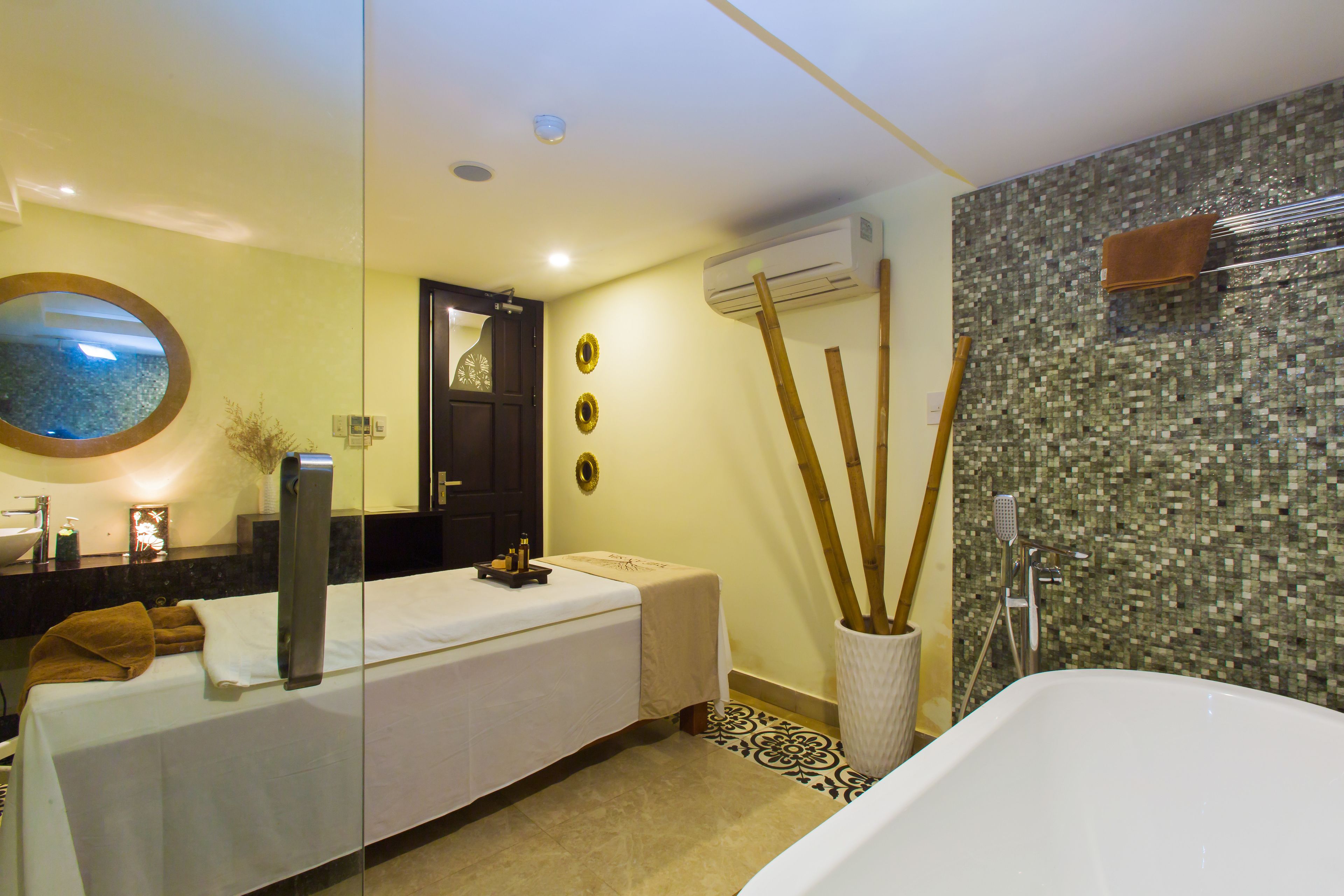 A modern bathroom with a glass partition, a large mirror, a massage table, a bathtub, and a mosaic wall.