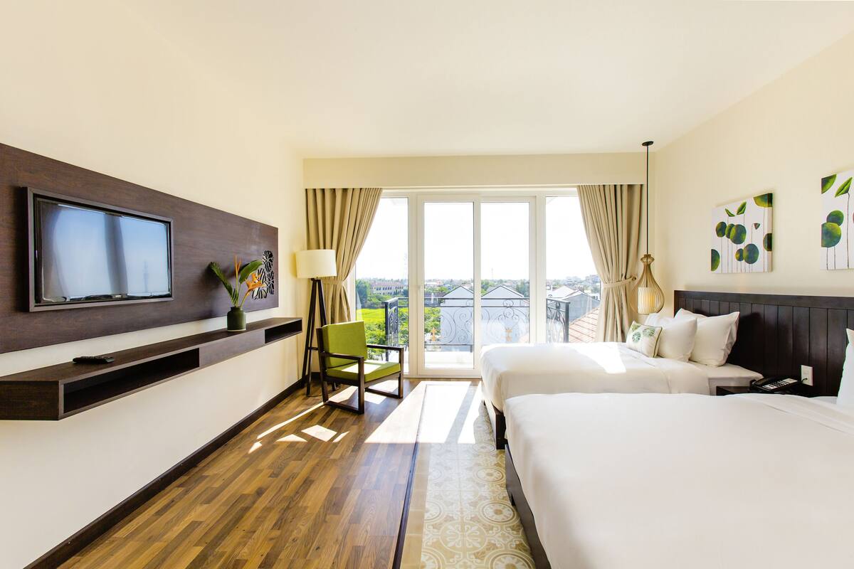 Une chambre d’hôtel avec un grand lit, un téléviseur fixé au mur, un bureau avec une lampe, une chaise et une vue sur la ville par la fenêtre.
