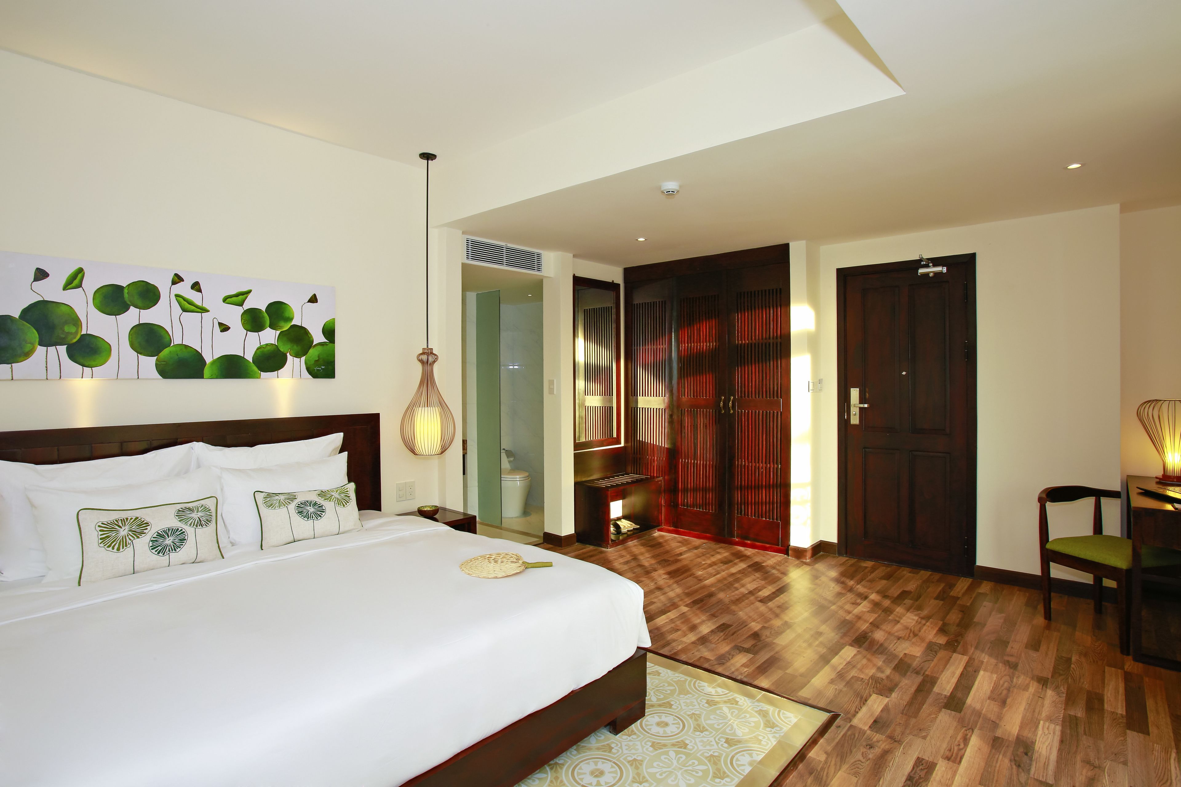 A hotel room with a large bed, a wooden headboard, a wall-mounted artwork, a bedside table, a nightstand, a lamp, and a door leading to another room.