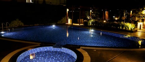 A nighttime swimming pool area with a circular hot tub, surrounded by well-lit plants and architectural features.
