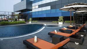 Un área de piscina en la azotea con sillones de descanso y sombrillas, rodeada por un edificio moderno con grandes ventanales.