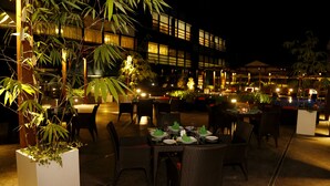Un área de comedor al aire libre con mesas preparadas para los invitados, rodeada de plantas en macetas y un edificio moderno al fondo.