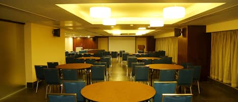 A conference room with multiple tables and chairs, equipped with ceiling lights and a projector screen.